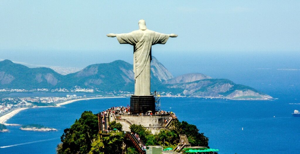 Foto do Cristo Redentor Visto de Costas com seus braços abertos. Ao fundo, a cidade do Rio e a praia.