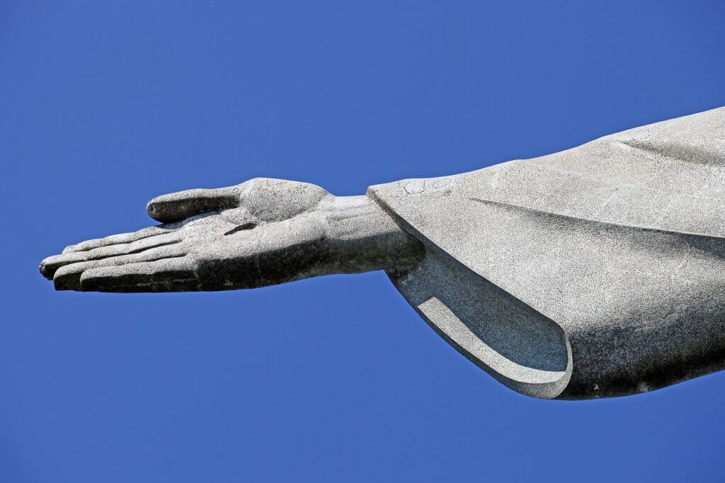 Braço direito do Cristo Redentor, a foto mostra apenas a mão furada do Cristo e parte da manga da roupa. Céu azul claro.