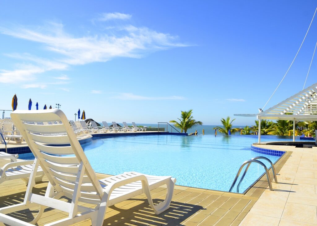 Foto de piscina de hotel, cadeira de praia a beira da piscina, ao fundo as águas do mar.